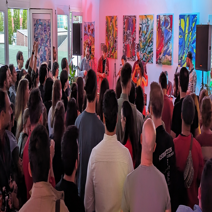 Foto von einem Konzert. Eine Band spielt auf einer kleinen Bühne. Im Hintergrund sind Gemälde zu sehen. Viele Zuschauer*innen sind im Raum versammelt.