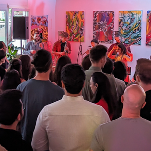 Foto von einem Konzert. Eine Band spielt auf einer kleinen Bühne. Im Hintergrund sind Gemälde zu sehen. Viele Zuschauer*innen sind im Raum versammelt.