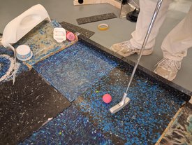 Person in heller Hose und weißen Schuhen spielt Minigolf auf einem Boden mit blauen und schwarzen Mustern und schlägt einen pinken Ball mit einem silbernen Putter