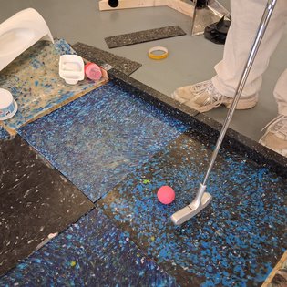 Person in heller Hose und weißen Schuhen spielt Minigolf auf einem Boden mit blauen und schwarzen Mustern und schlägt einen pinken Ball mit einem silbernen Putter