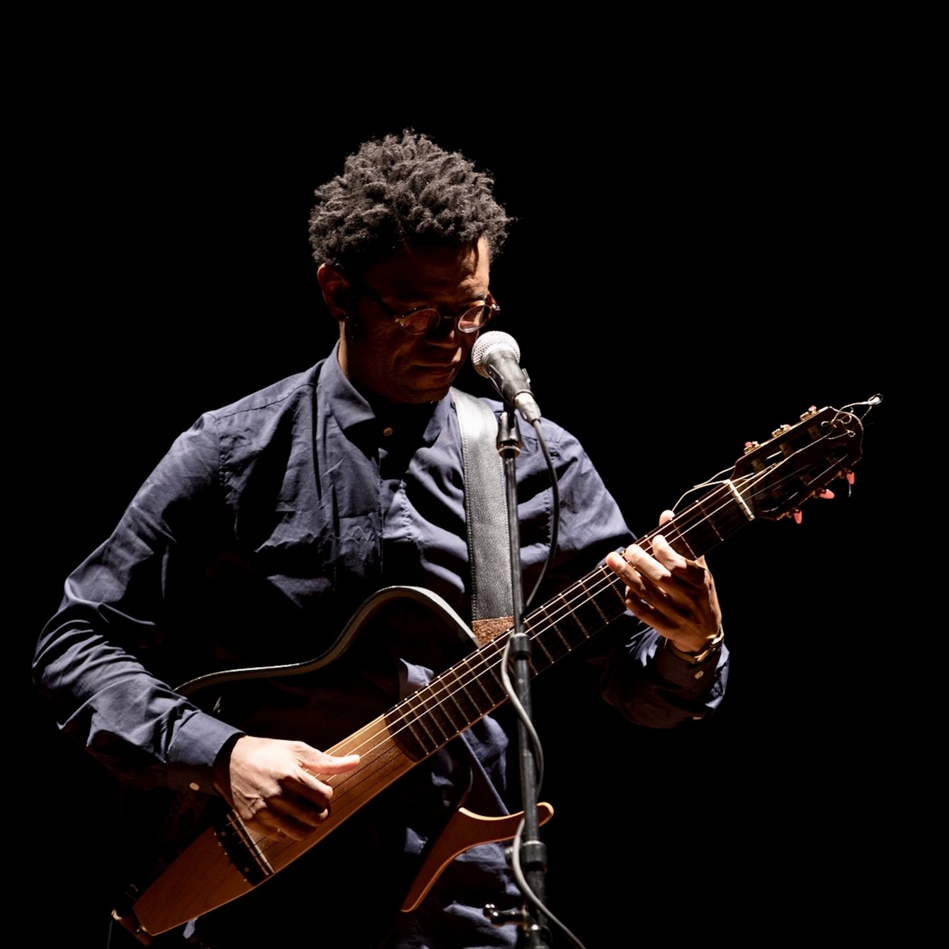 Neo Muyanga, bekleidet mit einem dunklen Hemd, spielt auf einer Gitarre. Er hat schwarze Haare und trägt eine Brille. Vor ihm steht ein Mikrofon.