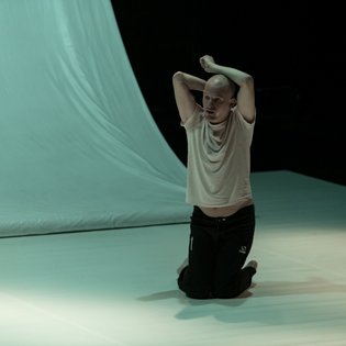Michael Turinsky auf der Bühne in einer Tanzpose auf den Knien. Er trägt ein helles Shirt und eine schwarze Hose.