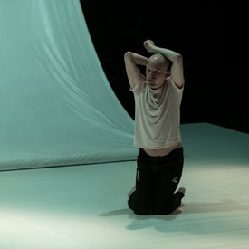 Michael Turinsky auf der Bühne in einer Tanzpose auf den Knien. Er trägt ein helles Shirt und eine schwarze Hose.