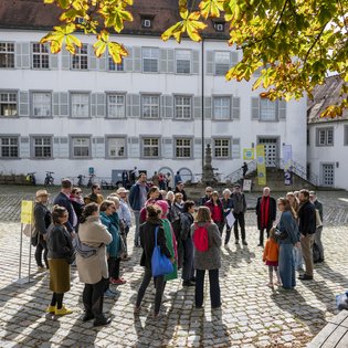 Die Gruppe der Teilnehmer*innen im Gespräch mit den Festival-Kurator*innen Herbordt/Mohren. Die Gruppe steht bei strahlendem Sonnenschein im Hof von Schloss Waldenbuch. Sie Sonne scheint. Herbstlich verfärbte Blätter eines Baumes ragen von oben in das Bild.