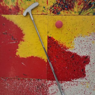Minigolf-Schläger mit weißem Griff lehnt auf buntem, abgenutztem Minigolf-Teppich neben einem roten Minigolfball