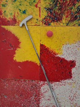 Minigolf-Schläger mit weißem Griff lehnt auf buntem, abgenutztem Minigolf-Teppich neben einem roten Minigolfball