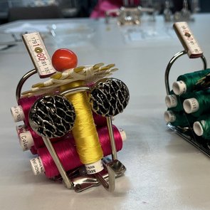 Zwei fantasievolle Figuren aus Metall, Garnrollen, Knöpfen und Maßbändern, die an Insekten erinnern, stehen auf einer glatten Oberfläche, dargestellt in einem kreisförmigen Ausschnitt, der wie ein Loch wirkt.
