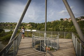 Zwei junge Menschen, möglicherweise ein Paar, stehen auf einer Aussichtsplatform und blicken ins Weite. Im Hintergrund ein Flusslauf mit Felsengärten und Weinanbau am Ufer.