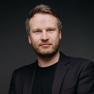 Mann mit kurzem Haar und Bart sitzt mit verschränkten Händen vor dunklem Hintergrund trägt schwarzen Blazer und schwarzen Pullover