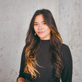 Junge Frau mit langen, gewellten, dunkelbraunen Haaren mit blonden Spitzen vor grauer Betonwand, trägt schwarzen Pullover und goldene Ohrringe.