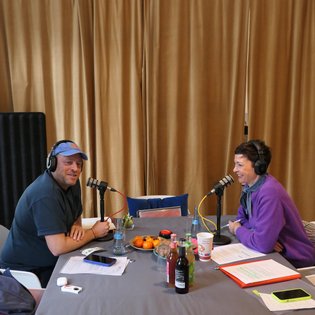 Zwei Personen, Kimsy von Reischach und Jacob Bilabel, sitzen an einem Tisch mit Mikrofonen, Getränken und Obst, vor einem goldfarbenen Vorhang.