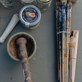 Foto von Mörser und Stößel, einer Dose mit der Aufschrift ‚Fichi Caramellati‘, mehreren Farbgläsern, benutzten Pinseln, einem Plastiklöffel und Glasbehältern.