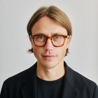 Ein junger Mann mit mittellangen, glatten, hellbraunen Haaren und blauen Augen blickt direkt in die Kamera. Er trägt eine braune Brille mit dickem Rahmen, ein schwarzes T-Shirt und ein schwarzes Jackett. Der Hintergrund ist neutral und hellgrau, was den Fokus auf die Person lenkt. Er hat einen ruhigen, freundlichen Gesichtsausdruck.