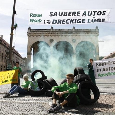 Bild einer Gruppe Aktivisten von Robn Wood auf einem städtischen Platz sitzend oder stehend. Zum Teil halten die Personen Banner in der Hand, zum Teil lehnen sie an Autoreifen, die auf dem Platz verteilt liegen.