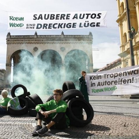 Bild einer Gruppe Aktivisten von Robn Wood auf einem städtischen Platz sitzend oder stehend. Zum Teil halten die Personen Banner in der Hand, zum Teil lehnen sie an Autoreifen, die auf dem Platz verteilt liegen.