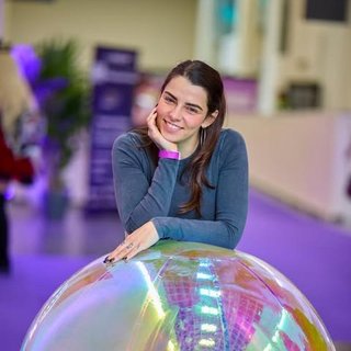 Eine junge Frau mit langen, braunen Haaren lehnt sich lächelnd auf eine große, glänzende, irisierende Kugel. Sie trägt ein langärmeliges, dunkelgraues Oberteil und ein rosa Armband. Die Kugel spiegelt farbenfrohe Lichtreflexionen in Lila-, Grün- und Goldtönen. Der Hintergrund ist unscharf, mit lila Bodenbelag und einem hellen, modernen Innenraum, vermutlich auf einer Messe oder Veranstaltung.