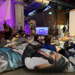 Teilnehmer sitzen auf bunten Sitzsäcken und schauen sich bei einem Symposium in Stuttgart eine Präsentation auf einem Bildschirm an.