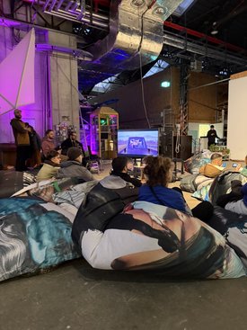 Teilnehmer sitzen auf bunten Sitzsäcken und schauen sich bei einem Symposium in Stuttgart eine Präsentation auf einem Bildschirm an.