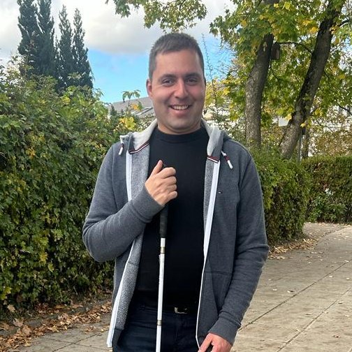 Lächelnder Mann mit Blindenstock steht auf einem von Bäumen gesäumten Gehweg mit Herbstlaub