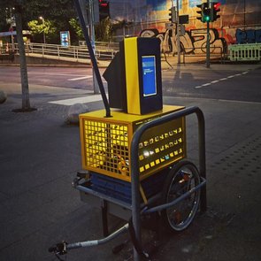  Eine gelb-schwarze Straßeninstallation mit Bildschirm und Leuchte steht auf einem Gehweg nahe einer Ampel. Auf dem Gerät steht „SOLUTIONS SOLUTIONS“. Im Hintergrund sind Graffiti zu sehen.