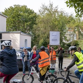 Während der Aufführung von Mauricio Kagels Stück »Eine Brise – Flüchtige Aktion für 111 Radfahrer« fahren mehrere Fahrradfahrer*innen an am Straßenrand stehendem Publikum vorbei. Ebenfalls am Straßenrand steht eine weiblich gelesene Person, die ein großes Schild mit der Aufforderung »ring, ring, ring, ring… « für die Radfahrer*innen in der Hand hält.