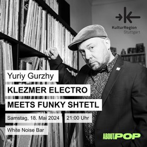 Yuriy Gurzhy lehnt an ein Regal voller Schalplatten. Bartträger Yuriy ist mit einer Kordmütze, einem gemusterten Hemd und einem dunklen Jacket bekleidet. 
