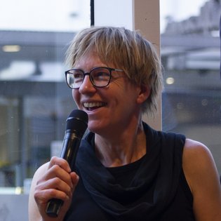 Person mit kurzen hellen Haaren und schwarzem ärmellosem Oberteil hält ein Mikrofon vor einem Fenster