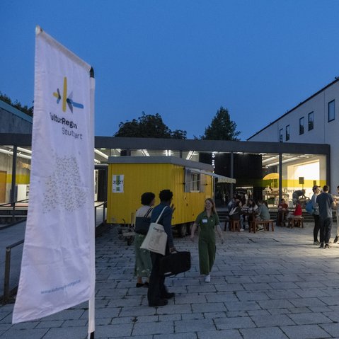 Eine Fahne und der gelbe Bauwagen der KulturRegion Stuttgart im Hof des Württembergischen Kunstvereins Stuttgart. Mehrere Personen sitzen an Tischen oder stehen frei im Hof. Es ist bereits dunkel und im Gebäude im Hintergrund brennt Licht.