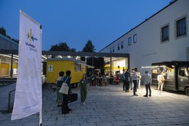Eine Fahne und der gelbe Bauwagen der KulturRegion Stuttgart im Hof des Württembergischen Kunstvereins Stuttgart. Mehrere Personen sitzen an Tischen oder stehen frei im Hof. Es ist bereits dunkel und im Gebäude im Hintergrund brennt Licht.