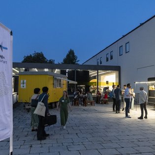 Eine Fahne und der gelbe Bauwagen der KulturRegion Stuttgart im Hof des Württembergischen Kunstvereins Stuttgart. Mehrere Personen sitzen an Tischen oder stehen frei im Hof. Es ist bereits dunkel und im Gebäude im Hintergrund brennt Licht.