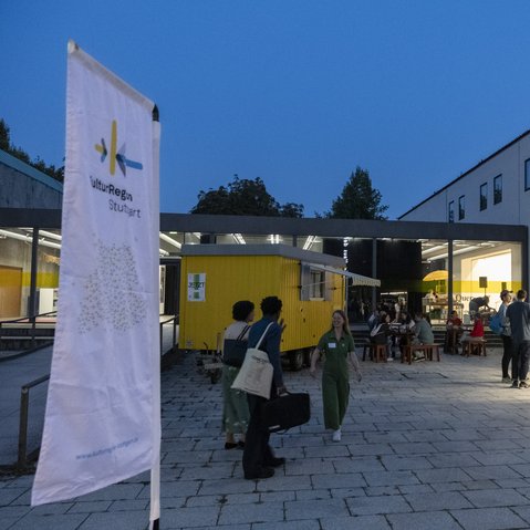 Eine Fahne und der gelbe Bauwagen der KulturRegion Stuttgart im Hof des Württembergischen Kunstvereins Stuttgart. Mehrere Personen sitzen an Tischen oder stehen frei im Hof. Es ist bereits dunkel und im Gebäude im Hintergrund brennt Licht.