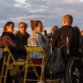 Rollstuhlnutzer Michael Turinsky, Anke Hofmann und Lucie Schroeder von hinten. Anke Hofmann und Lucie Schröder sitzen auf gelben Stühlen. Anke Hofmann spricht in ein Mikrofon. Abendsonne beleuchtet die Szene.