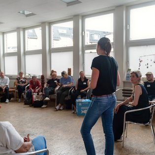 Personengruppe sitzt in einem Kreis in einem hellen Raum mit großen Fenstern. Eine Person steht und spricht offenbar zur Gruppe. Der Raum ist mit modernen Möbeln, einem Korkboden und verschiedenen Materialien ausgestattet, darunter ein Karton mit der Zahl ‚14‘ und mehrere Taschen. Die Szene deutet auf ein Treffen, einen Workshop oder eine Diskussionsrunde hin.