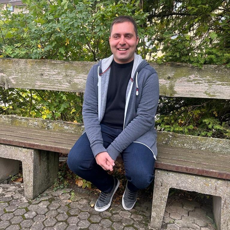 Bild von Matthias Nagel auf einer Holzbank sitzend. Er trägt einen blauen Hoodie über einem schwarzen Shirt. Dazu Jeans. Matthias Nagel hat kurzes, braunes Haar und lächelt freundlich in die Kamera.