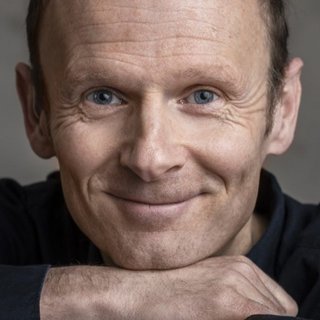 Jochen Weber hat kurze Haare und lächelt freundlich in die Kamera. Er stützt sein Kinn auf seine überkreuzten Arme und trägt ein dunkles Hemd. Der Hintergrund ist neutral und unscharf, sodass der Fokus auf seinem Gesicht liegt.