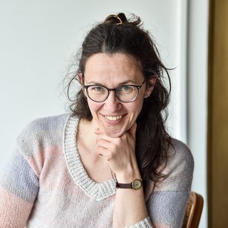 Porträt von Boglárka Pap. Sie hat dunkle, lange Haare, Brille und trägt einen gestrickten Pullover in Pastellfarben. Sie sitzt lächelnd mit einer Hand am Kinn vor einem hellen Hintergrund.