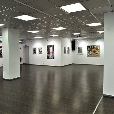 Innenansicht einer modernen Kunstgalerie mit weiß gestrichenen Wänden, dunklem Holzboden und Rasterdeckenbeleuchtung. An den Wänden hängen verschiedene Gemälde und Fotografien, die Szenen mit Menschen darstellen. Die Galerie ist geräumig, gut beleuchtet und wirkt ruhig und einladend.
