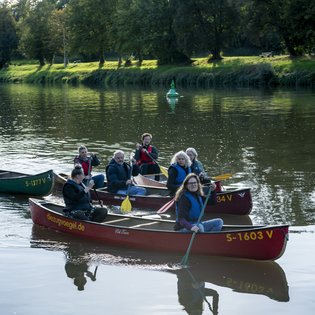 Vier mit Personen besetzte Kanus, die nah aneinander auf einem Gewässer fahren.