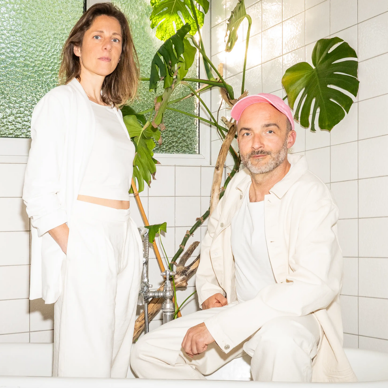 Bild von Melanie und Markus Niessner in einer Badewanne in einem weiß-gekachelten Raum mit einer großen Pflanze im Hintergrund. Beide sind weiß gekleidet, Markus Niessner trägt dazu eine rosa Baseballkappe. Melanie Niessner steht in der Badewanne, Markus Niessner sitzt neben ihr auf dem Rand der Badewanne.