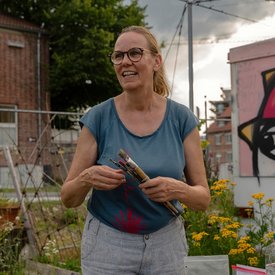 Barbara Karsch-Chaïeb in blauem T-Shirt mit rotem Blattmuster steht in einem urbanen Garten und hält mehrere Pinsel. Im Hintergrund sind gelbe Blumen, ein Zaun, ein Kunstwerk und ein Gebäude zu sehen.