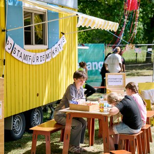 vor einem gelben Bauwagen hängen bunte Wimpel im Baum, im Vordergrund ist eine Person die einen Flyer liest und Menschen sitzen auf Liegestühlen.