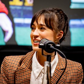 Nahaufnahme von Rocío Alemán vor einem Mikrofon. Sie hat dunkle Haare, die am Hinterkopf hochgesteckt sind, trägt eine weiße Bluse und ein kariertes Jacket und lächelt einem Gesprächspartner zu.