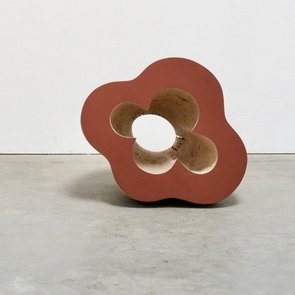 Eine braune Skulptur, eventuell aus Holz, steht auf einem Betonboden vor einer weißen Wand.