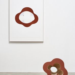 Eine braune Skulptur, eventuell aus Holz, steht auf einem Betonboden vor einer weißen Wand.