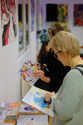Zwei weiblich gelesene Personen schauen sich Broschüren an. In den Räumlichkeiten hängen Kunstwerke an der Wand.