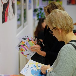 Zwei weiblich gelesene Personen schauen sich Broschüren an. In den Räumlichkeiten hängen Kunstwerke an der Wand.