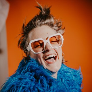 Person mit hochgestecktem Haar trägt eine blaue, flauschige Jacke vor orangefarbener Wand