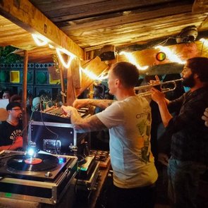 Ein DJ bedient bei einer Veranstaltung im Freien das DJ-Pult. Rechts vom DJ spielt eine Person auf einer Trompete. Im Hintergrund sind tanzende Menschen und das Soundsystem zu sehen.