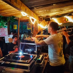Ein DJ bedient bei einer Veranstaltung im Freien das DJ-Pult. Rechts vom DJ spielt eine Person auf einer Trompete. Im Hintergrund sind tanzende Menschen und das Soundsystem zu sehen.