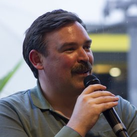 Mann mit dunklem Haar und grünem Poloshirt hält ein Mikrofon und spricht